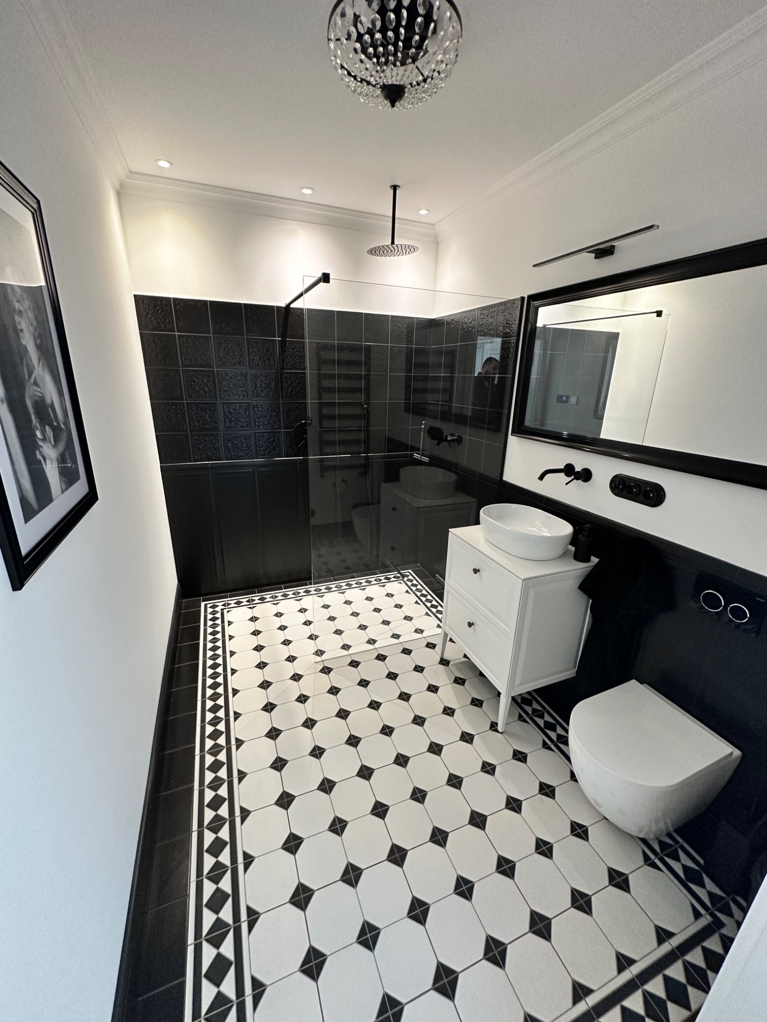 Elegancka łazienka w czerni i bieli z geometryczną podłogą, kabiną prysznicową typu walk-in, białą szafką z umywalką nablatową i wiszącą toaletą.