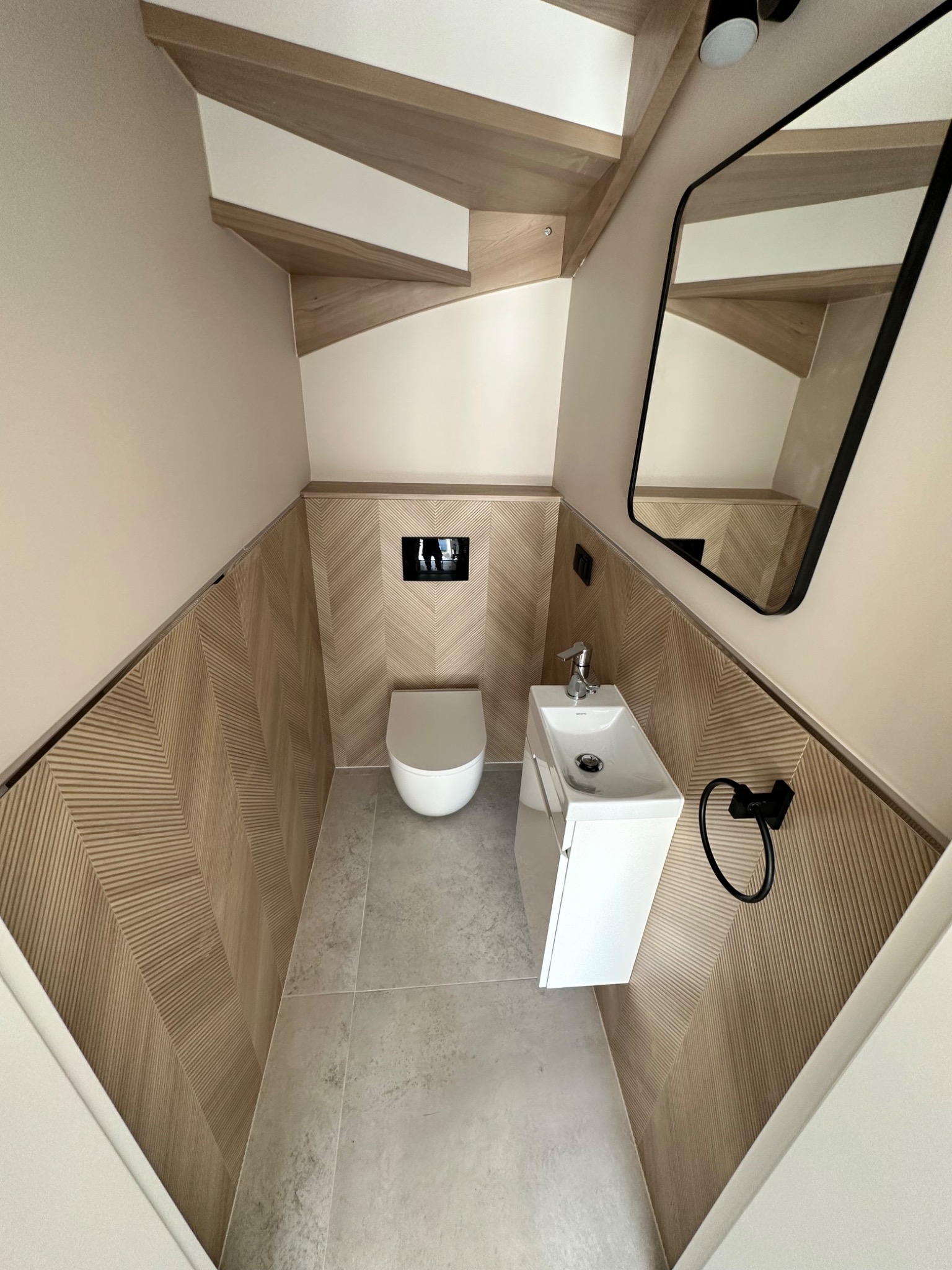 Niewielka łazienka pod schodami z toaletą podwieszaną, umywalką nablatową na szafce, lustrem w czarnej ramie i panelami ściennymi w jodełkę.