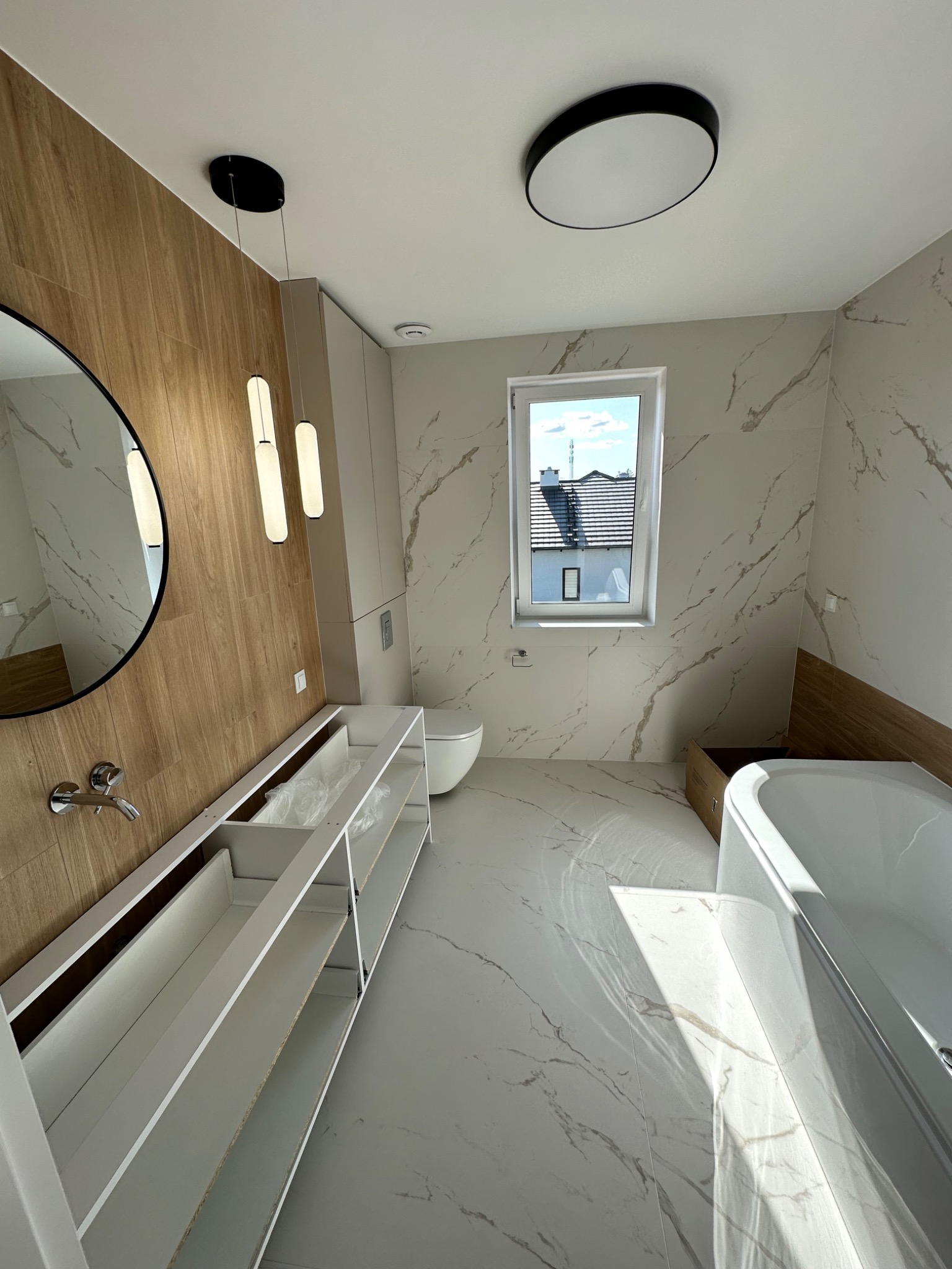 Nowoczesna łazienka z marmurowymi płytkami, drewnianą okładziną, okrągłym lustrem, wiszącymi lampami i częściowo zamontowaną szafką pod umywalkę.