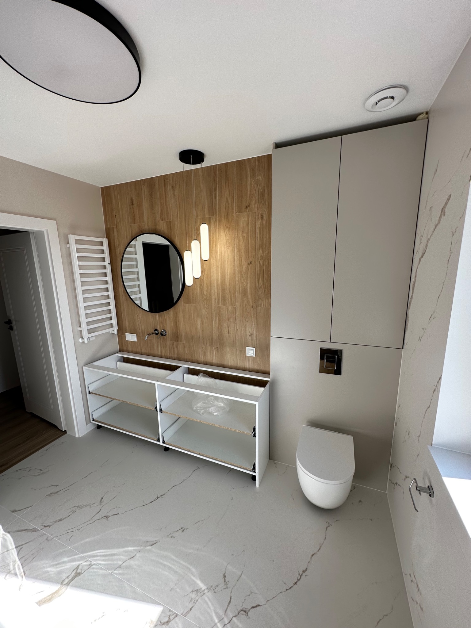 Nowoczesna łazienka w trakcie montażu: biała szafka pod umywalkę bez frontów, okrągłe lustro z trzema podłużnymi lampami, wisząca toaleta i marmurowe płytki podłogowe.