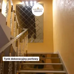 Perłowy tynk dekoracyjny na ścianie klatki schodowej, odbijający światło w lustrzanej mozaice i lśniących stopniach schodów z balustradą z przezroczystego tworzywa ze złotymi elementami.