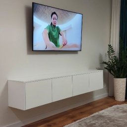 METER DECOR VADYM TYMOSEVYCH - Nowoczesny salon z podwieszaną białą szafką RTV, na ścianie telewizor wyświetlający masażystkę, drewniana podłoga i okrągły dywan z frędzlami.