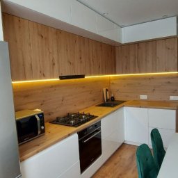 METER DECOR VADYM TYMOSEVYCH - Nowoczesna kuchnia w kształcie litery L z białymi frontami dolnych szafek i drewnopodobnymi górnymi szafkami, podświetlona listwą LED, z czarną płytą grzewczą i piekarnikiem, widoczny fragment...