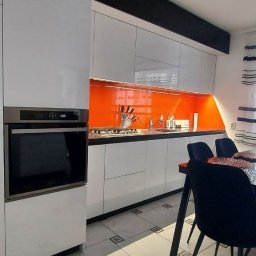METER DECOR VADYM TYMOSEVYCH - Nowoczesna biała kuchnia z pomarańczowym panelem nad blatem i zabudowanym piekarnikiem, widoczne krzesła przy stole.