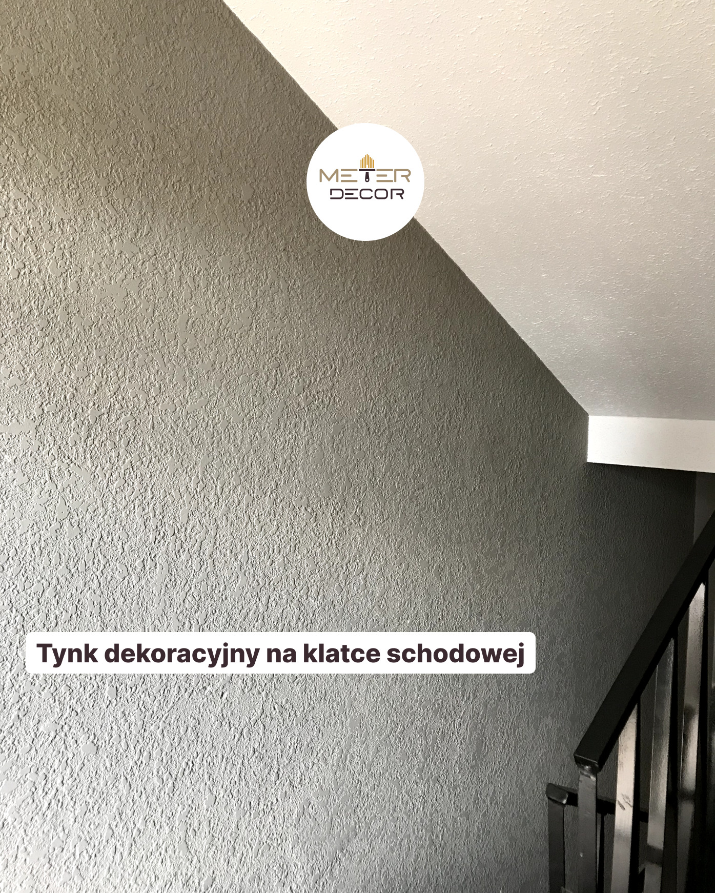 Szara ściana na klatce schodowej pokryta dekoracyjnym tynkiem o wyraźnej fakturze, widoczny fragment białego sufitu i czarna balustrada schodów.