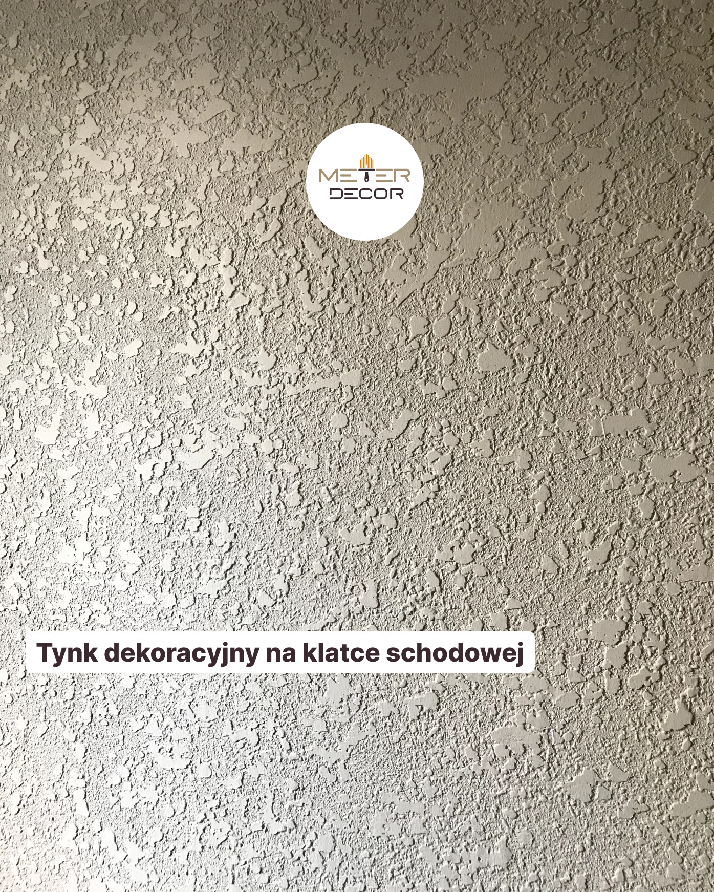 Szary tynk dekoracyjny o nieregularnej fakturze na ścianie klatki schodowej, z widocznym logo firmy w górnej części zdjęcia.