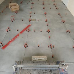 RemPejs - Układanie szarych płytek podłogowych z użyciem systemu poziomowania; widoczna poziomica i przecinarka do płytek.