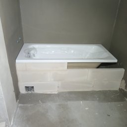 RemPejs - Nowa, biała wanna akrylowa, jeszcze w folii ochronnej, osadzona na wymurowanym cokole z bloczków, widoczne przyłącze kanalizacyjne w ścianie, surowe ściany łazienki przed wykończeniem.