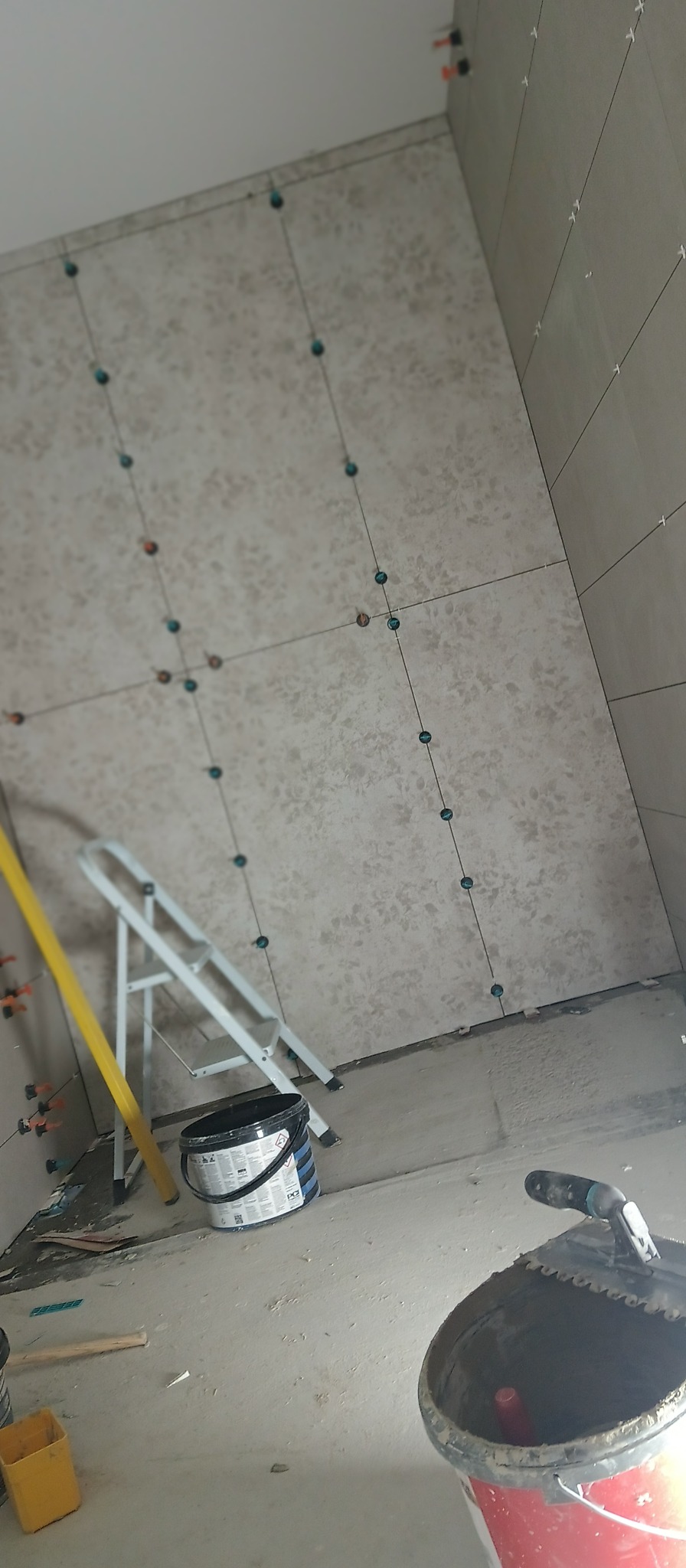 Ściana w trakcie układania płytek w Kłyżowie, widoczne poziomice, drabina i wiadro z zaprawą, płytki z delikatnym wzorem i system poziomowania płytek.