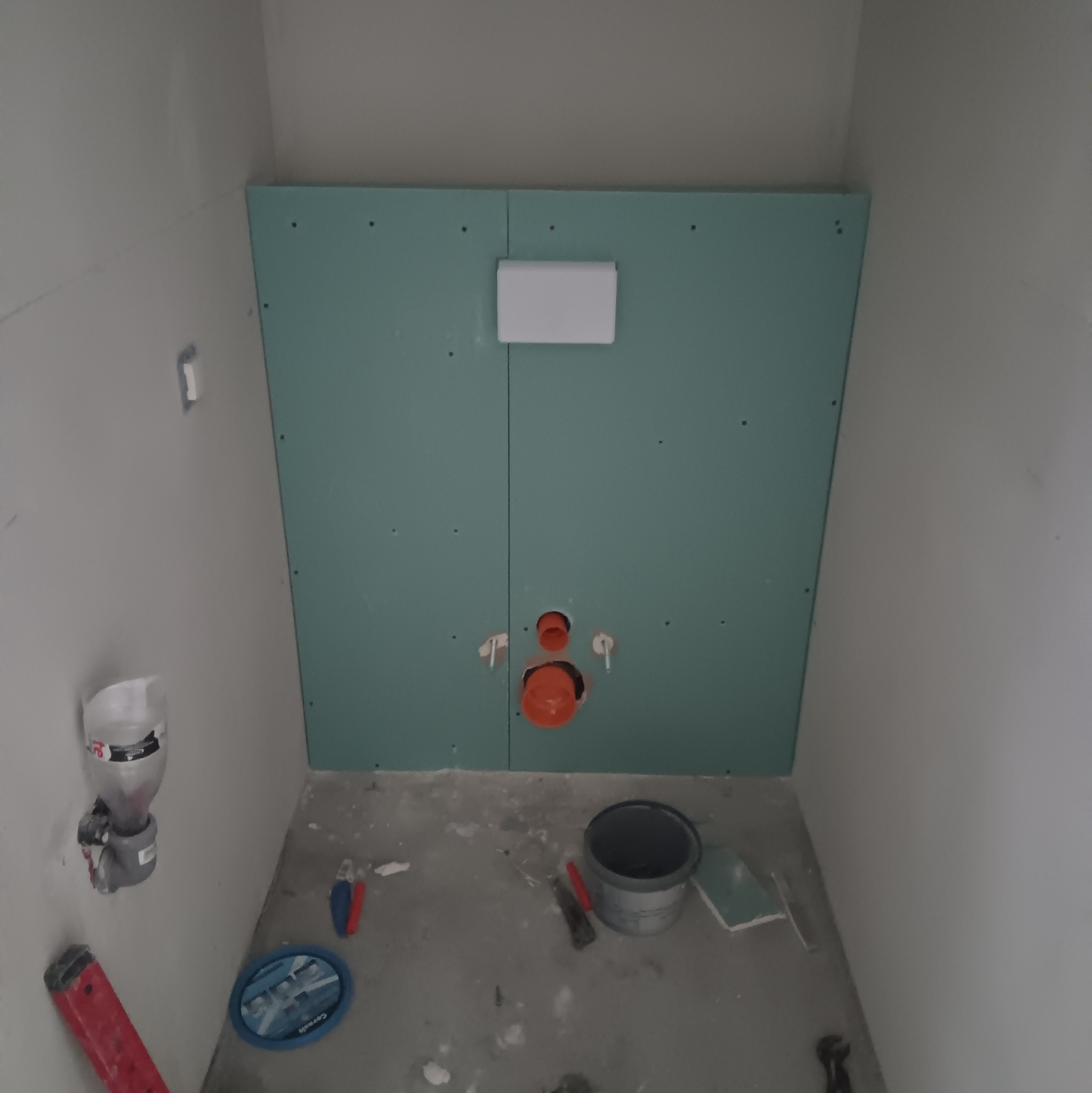 Wnętrze łazienki w trakcie prac: widoczne płyty kartonowo-gipsowe na ścianie z przygotowanymi otworami na instalację sanitarną, plastikowy zbiornik na ścianie, wiadro, poziomica i inne narzędzia...