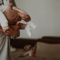 Paulina Macieląg Fotografia - Dłonie ojca trzymające noworodka w białym ubranku i skarpetkach, widoczny zegarek na nadgarstku mężczyzny.