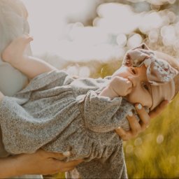 Paulina Macieląg Fotografia - Matka trzyma na rękach niemowlę w szarej sukience i opasce z kokardą na tle rozmytej zieleni