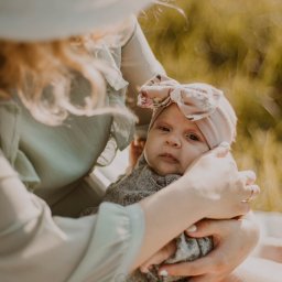 Paulina Macieląg Fotografia - Matka w pastelowej sukience trzyma na kolanach niemowlę z różową opaską na głowie z kokardą w kwiaty, na tle rozmytego zielonego pola.