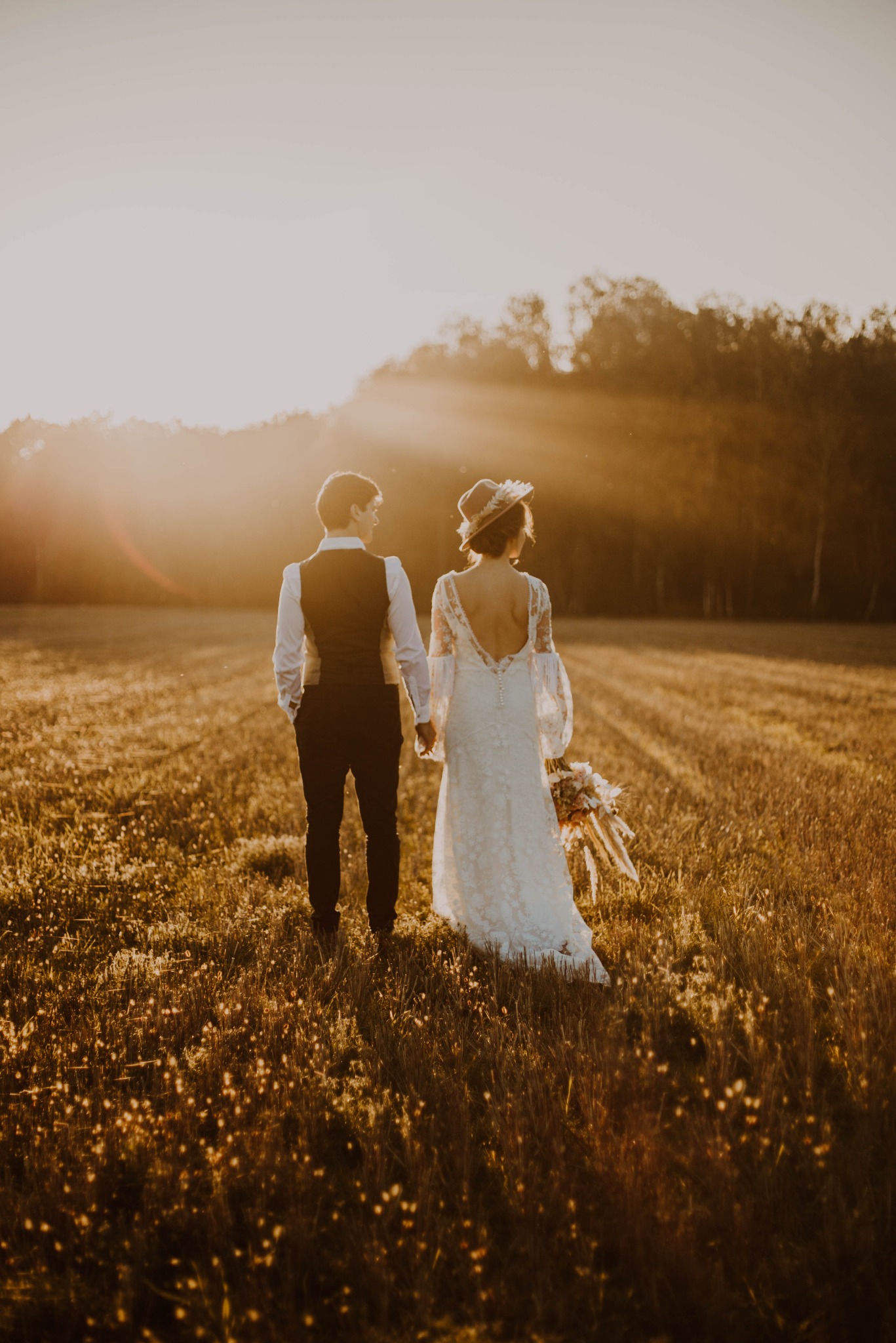 Para młoda, trzymająca się za ręce, spaceruje po zalanym złotym światłem polu trawy podczas zachodu słońca. Panna młoda w koronkowej sukni z odkrytymi plecami i kapeluszu, pan młody w kamizelce...