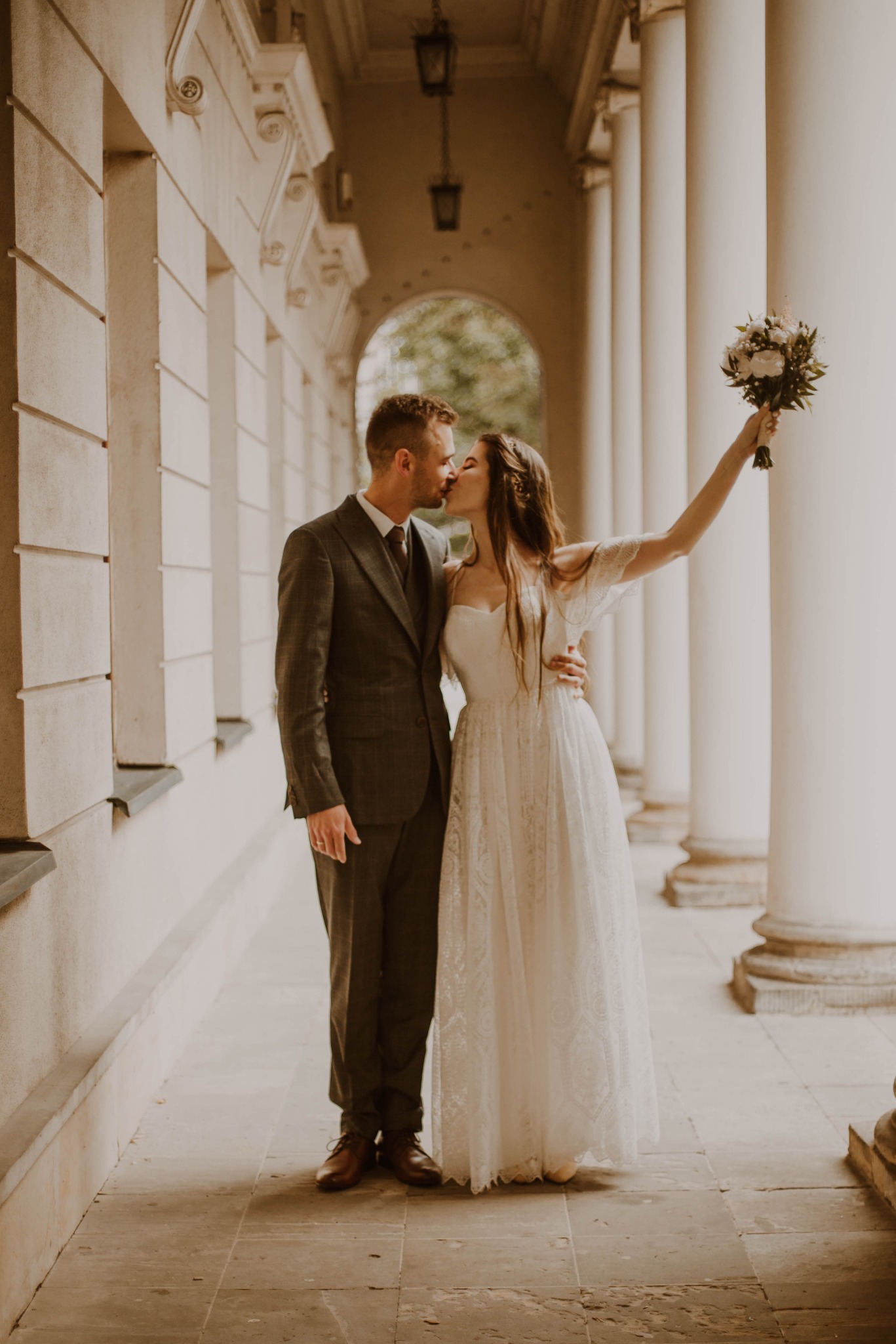 Pocałunek pary młodej w kolumnadzie, panna młoda z uniesionym bukietem w ręku, suknia z koronkowymi wstawkami, garnitur w kratę.