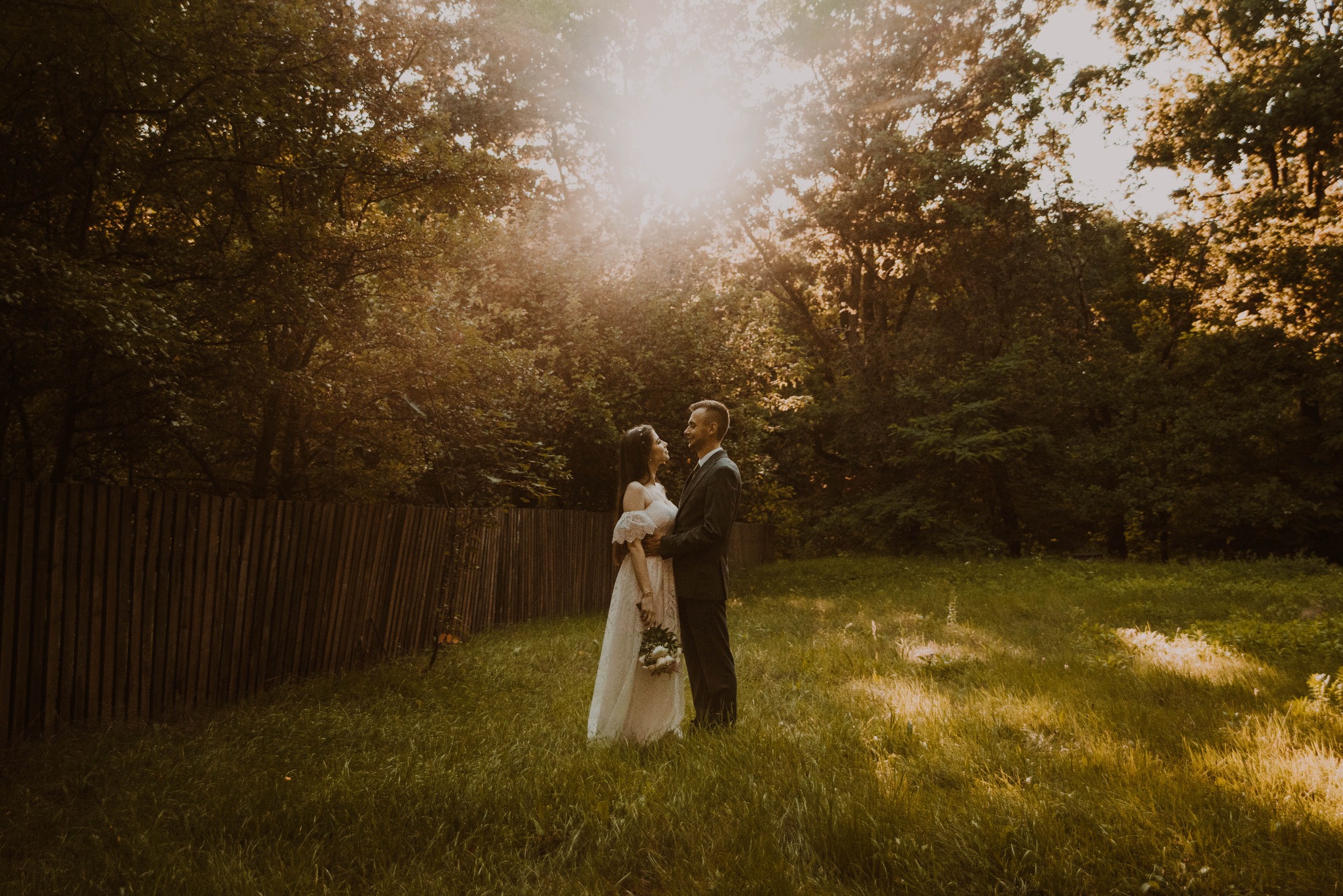 Para młoda w objęciach na łące w promieniach słońca, ona w sukni z bukietem, on w garniturze, drewniany płot w tle.