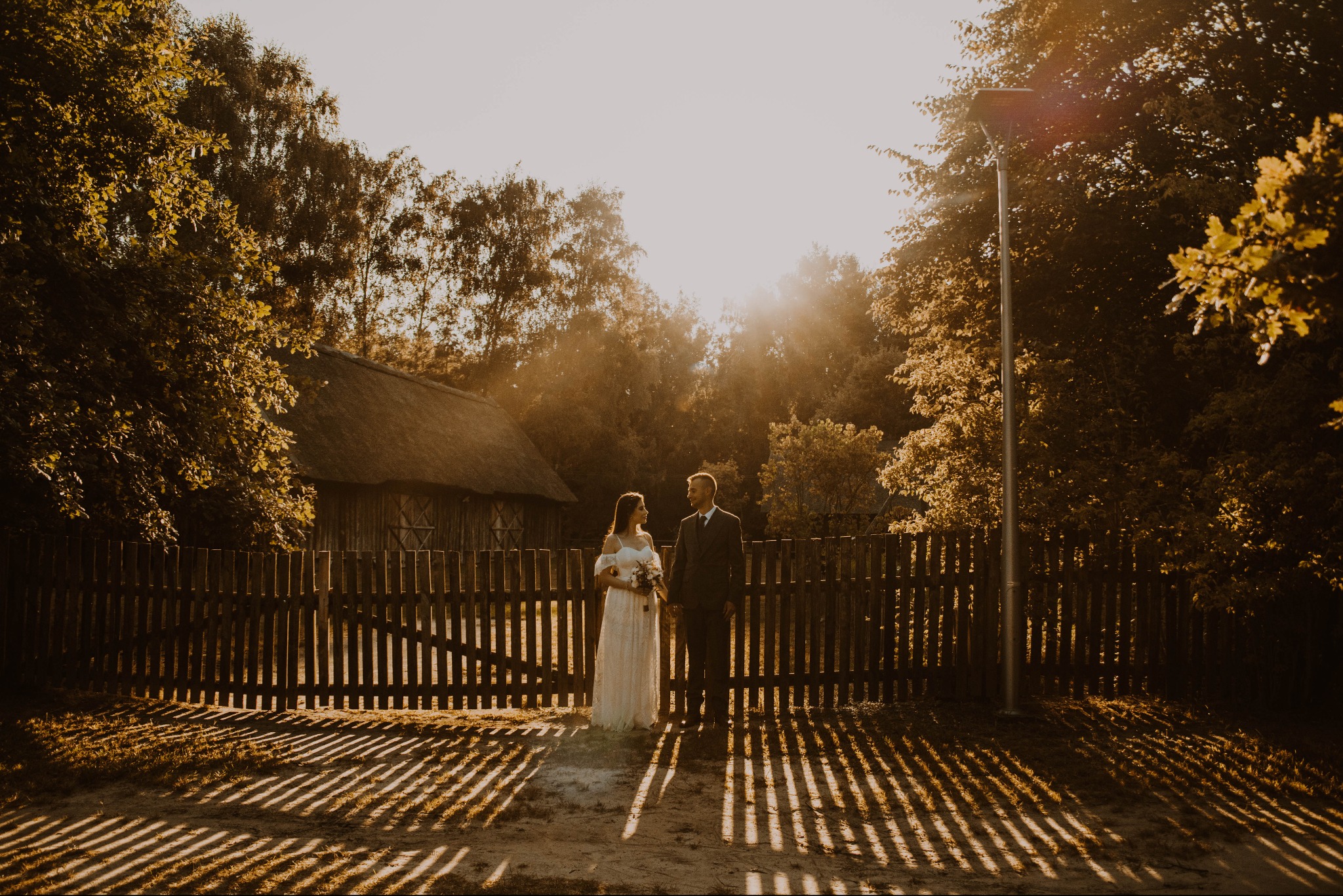 Para młoda w strojach ślubnych stoi przed drewnianym płotem w wiejskim otoczeniu, w tle widoczny budynek z dachem krytym strzechą, promienie zachodzącego słońca tworzą długie cienie.