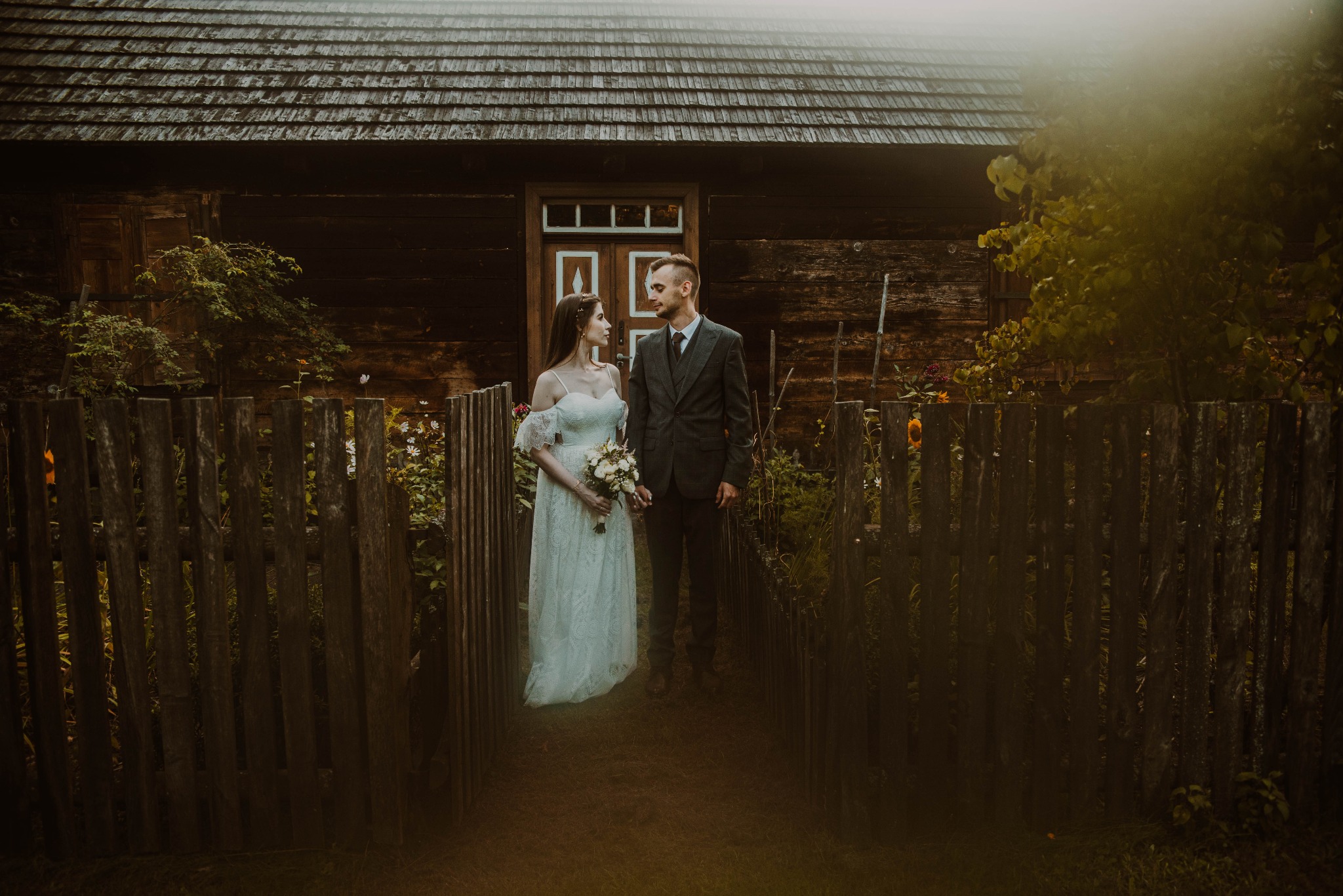 Para młoda w rustykalnej scenerii: panna młoda w koronkowej sukni i pan młody w tweedowym garniturze, stoją na tle starej, drewnianej chaty z drewnianym płotem, patrząc na siebie z czułością.