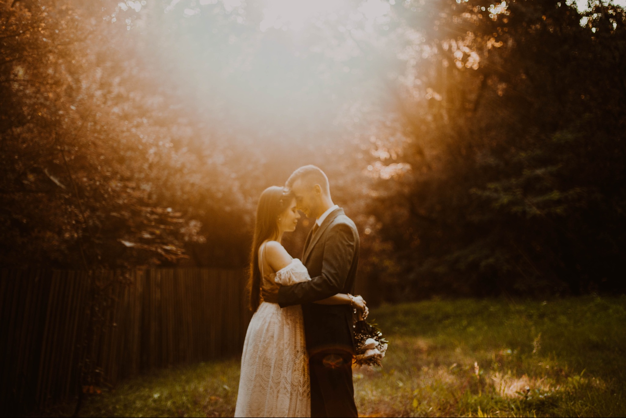 Para młoda przytulona w lesie w promieniach zachodzącego słońca, panna młoda w koronkowej sukni, pan młody w ciemnym garniturze trzyma bukiet białych róż.