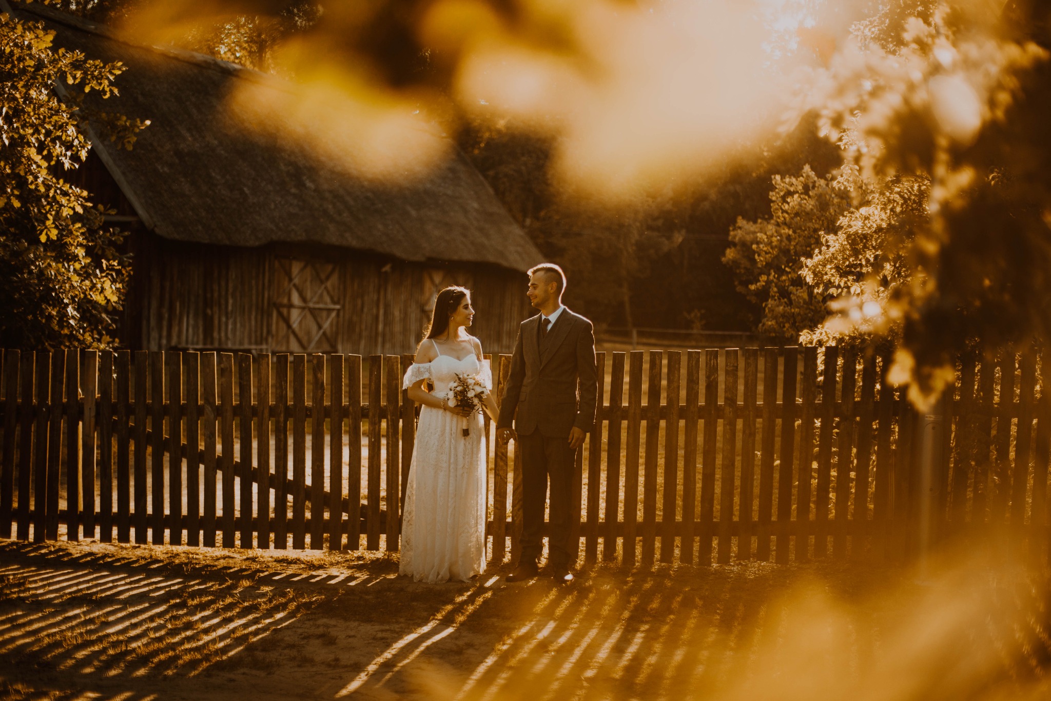 Para młoda w rustykalnej scenerii: panna młoda w koronkowej sukni z bukietem, pan młody w garniturze, stoją przy drewnianym płocie na tle starej chaty w promieniach zachodzącego słońca.