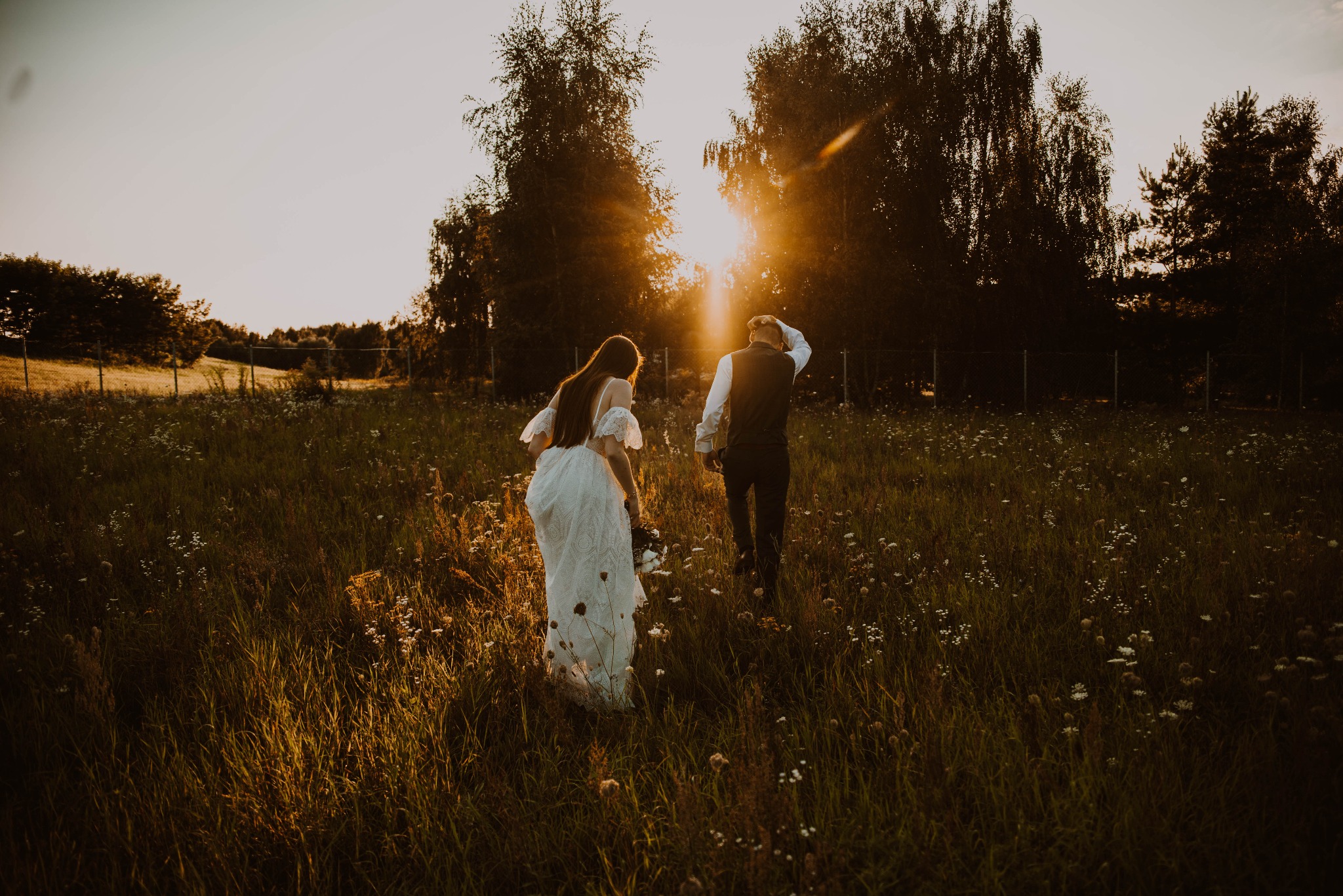 Para młoda, odwrócona tyłem, idzie przez łąkę w promieniach zachodzącego słońca, on w kamizelce, ona w koronkowej sukni, trzyma bukiet.