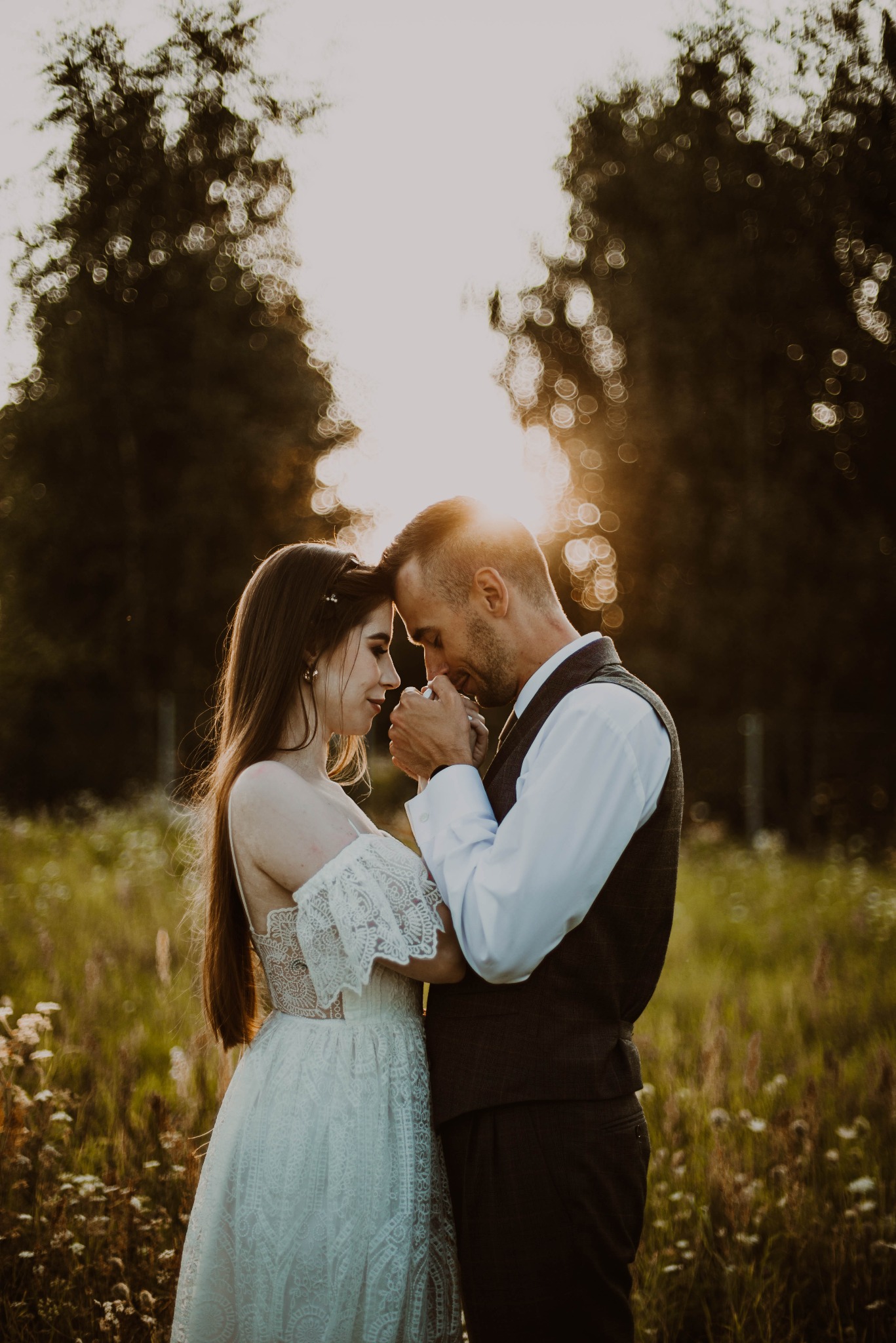 Para młoda w rustykalnej sesji ślubnej na łące o zachodzie słońca, panna młoda w koronkowej sukni z odkrytymi ramionami, pan młody w kamizelce, trzymają się za ręce.