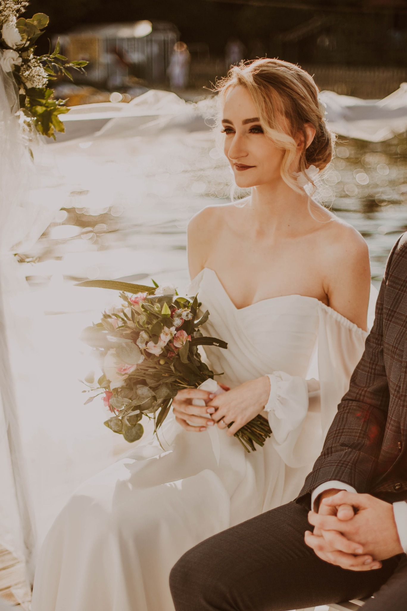 Panna młoda w białej sukni z odkrytymi ramionami, trzyma bukiet kwiatów, siedzi obok mężczyzny w kraciastej marynarce na tle jeziora w słoneczny dzień.