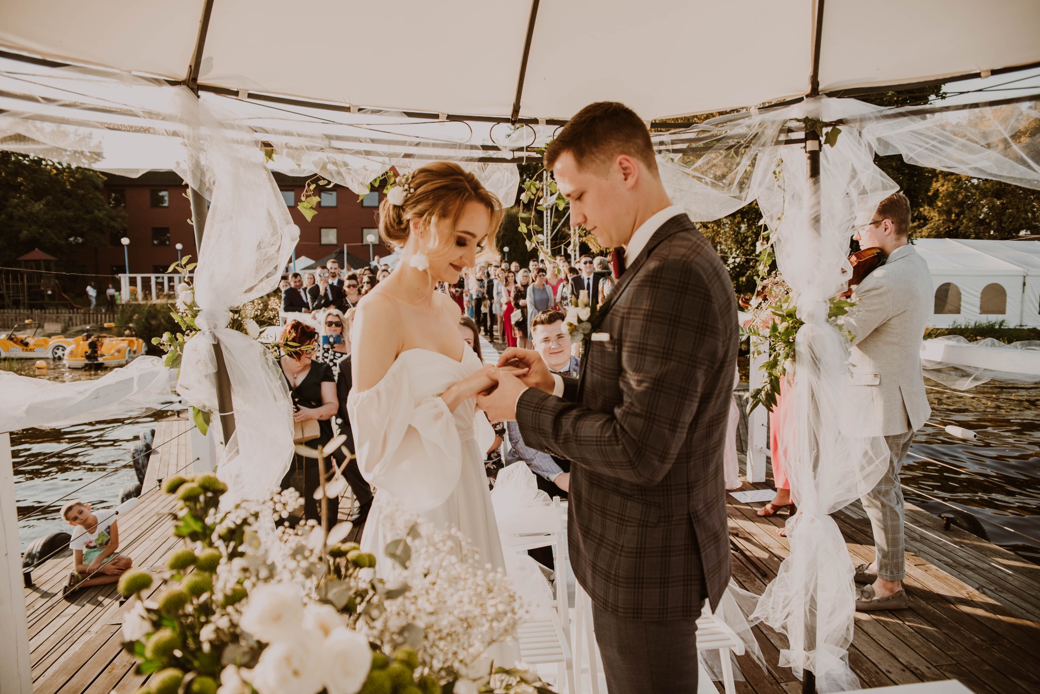 Wymiana obrączek podczas ceremonii ślubnej w Niedrzwicy Dużej, na drewnianym pomoście nad wodą, w tle goście i muzyk grający na skrzypcach.