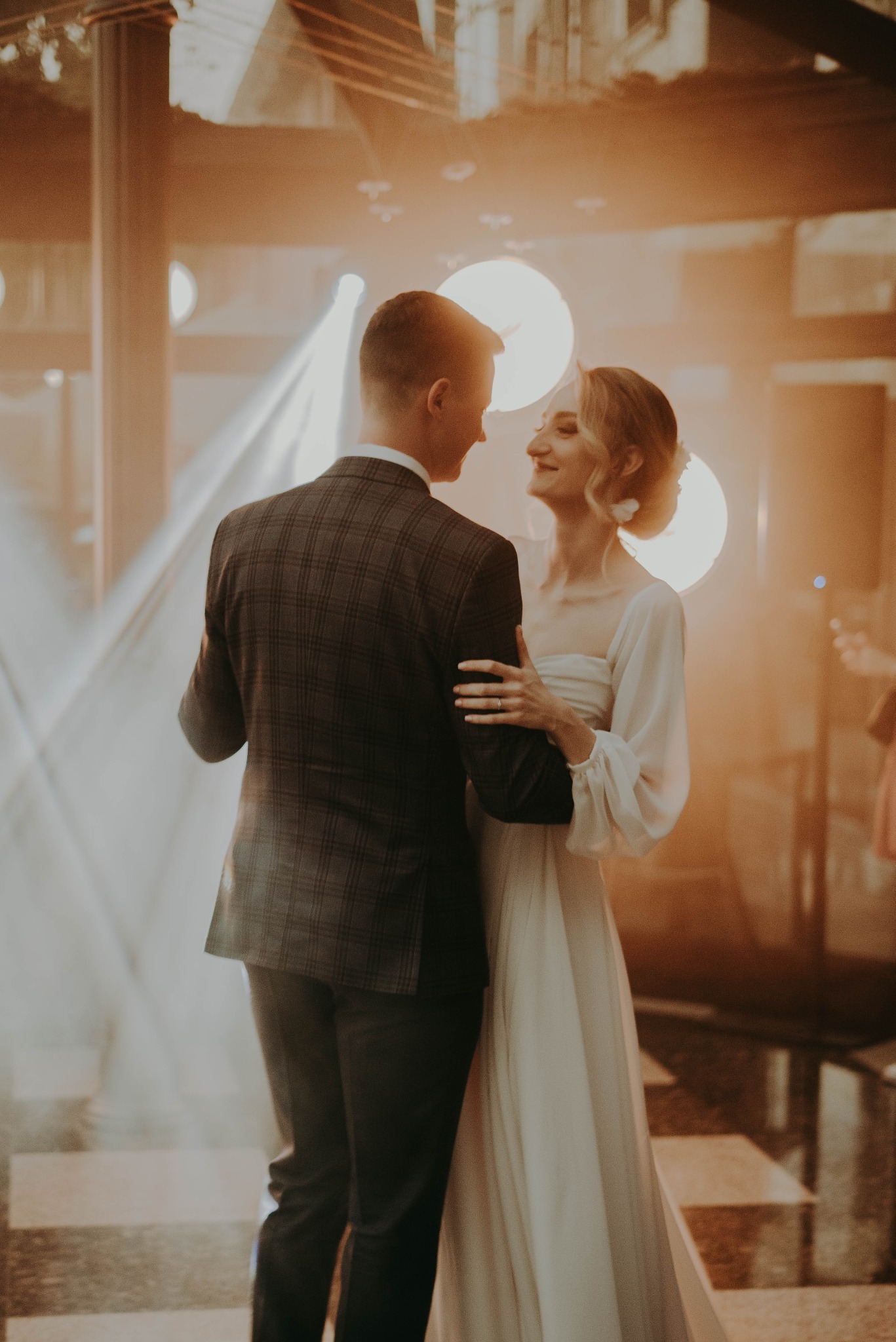 Para młoda w tańcu, ona w białej sukni z długimi rękawami, on w brązowym garniturze w kratę, oświetleni ciepłym światłem reflektorów, uśmiechnięci do siebie.