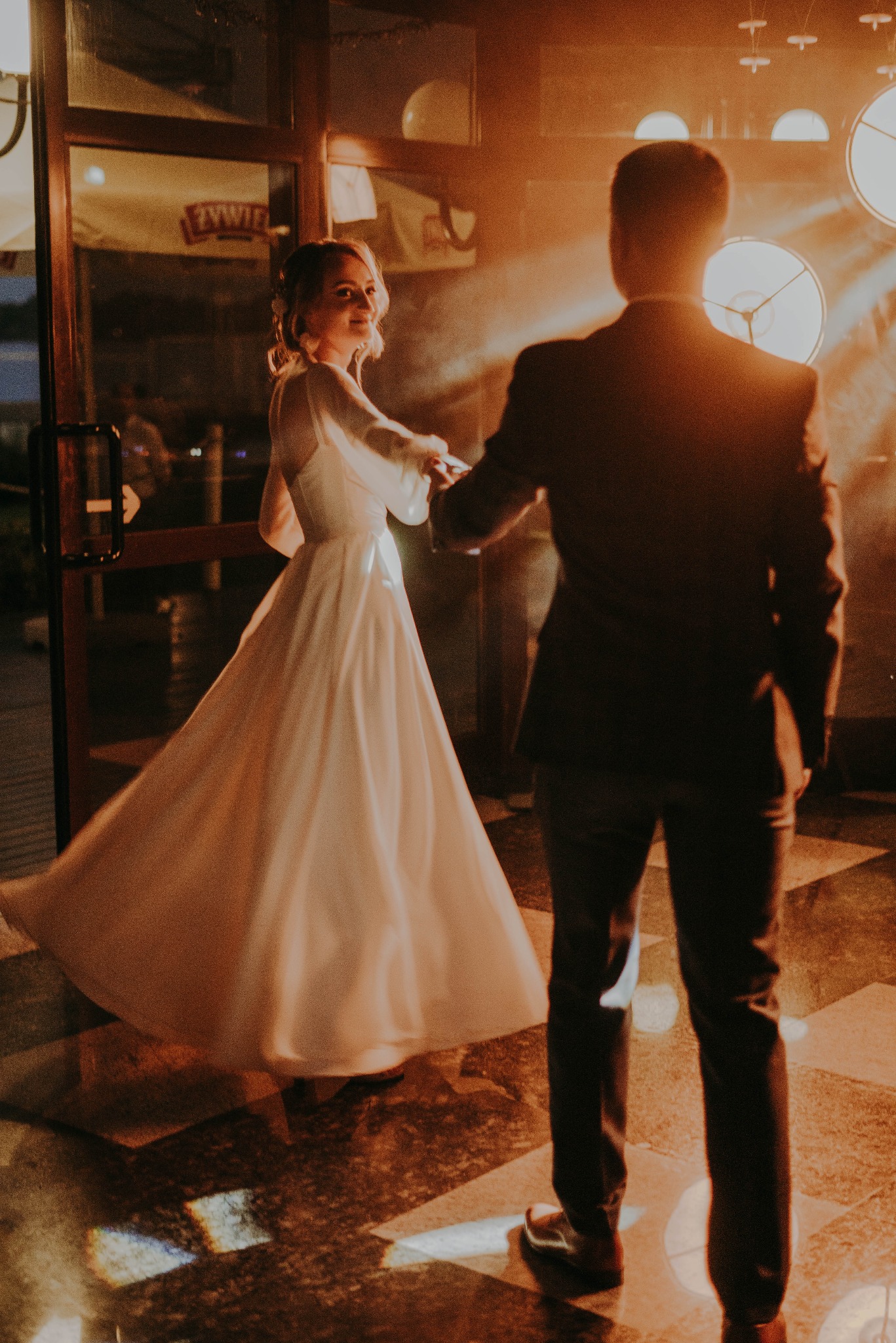 Panna młoda w białej sukni, odwrócona i uśmiechnięta, trzyma dłoń pana młodego w ciemnym garniturze na tle ciepłego oświetlenia i dekoracji sali.