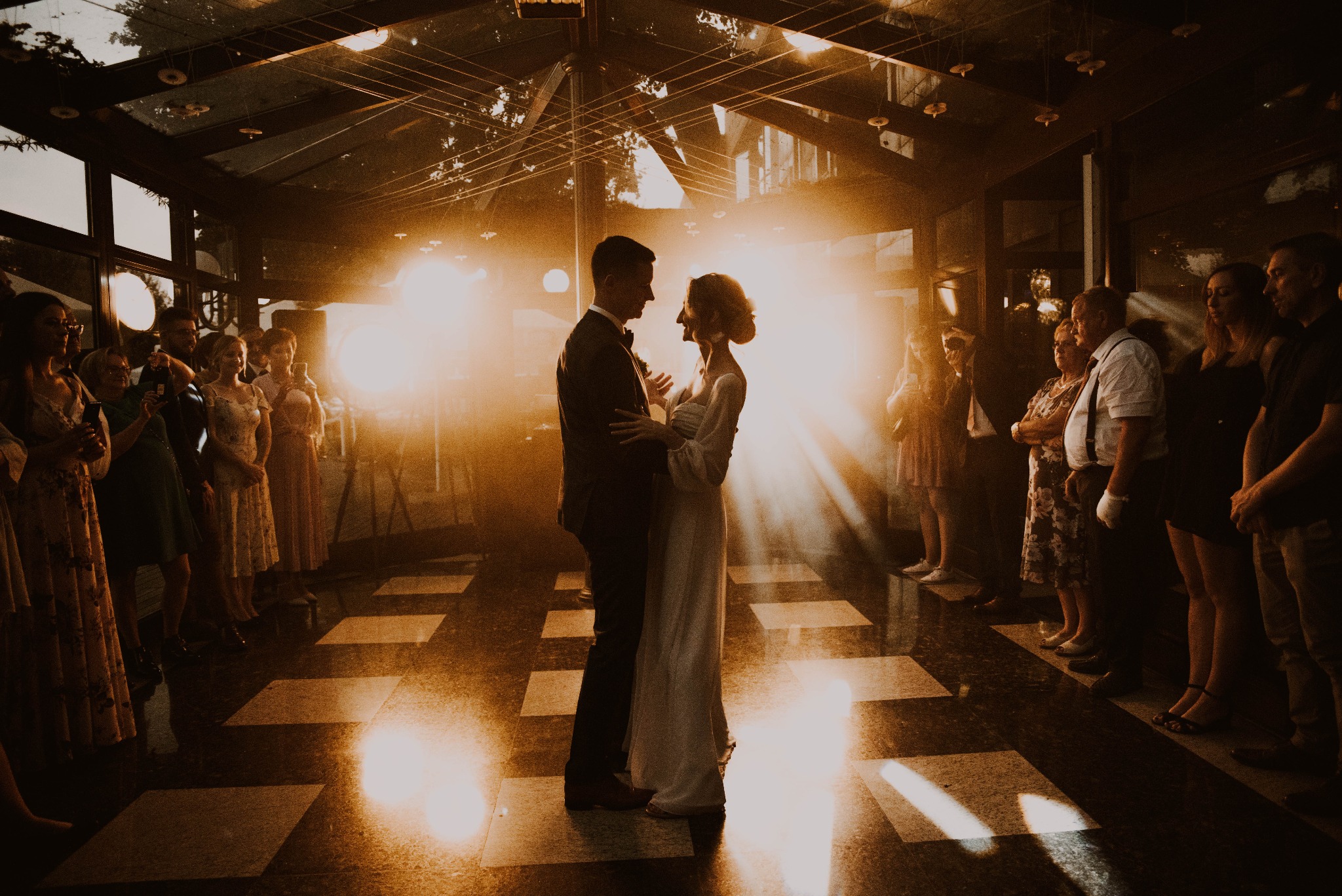 Para młoda w objęciach na parkiecie podczas reportażu ślubnego, otoczona gośćmi weselnymi w klimatycznym, oświetlonym wnętrzu.