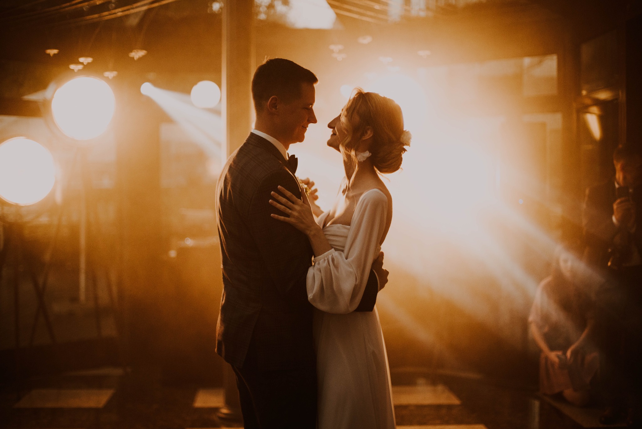 Para młoda w tańcu podczas reportażu ślubnego, oświetlona ciepłym światłem reflektorów, on w brązowym garniturze w kratę, ona w prostej białej sukni z bufkami, z kwiatami we włosach.