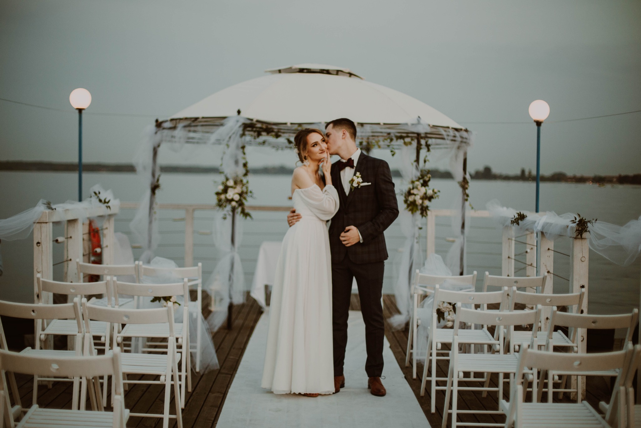 Para młoda na pomoście, mężczyzna całuje kobietę w policzek, w tle dekoracje ślubne i jezioro.