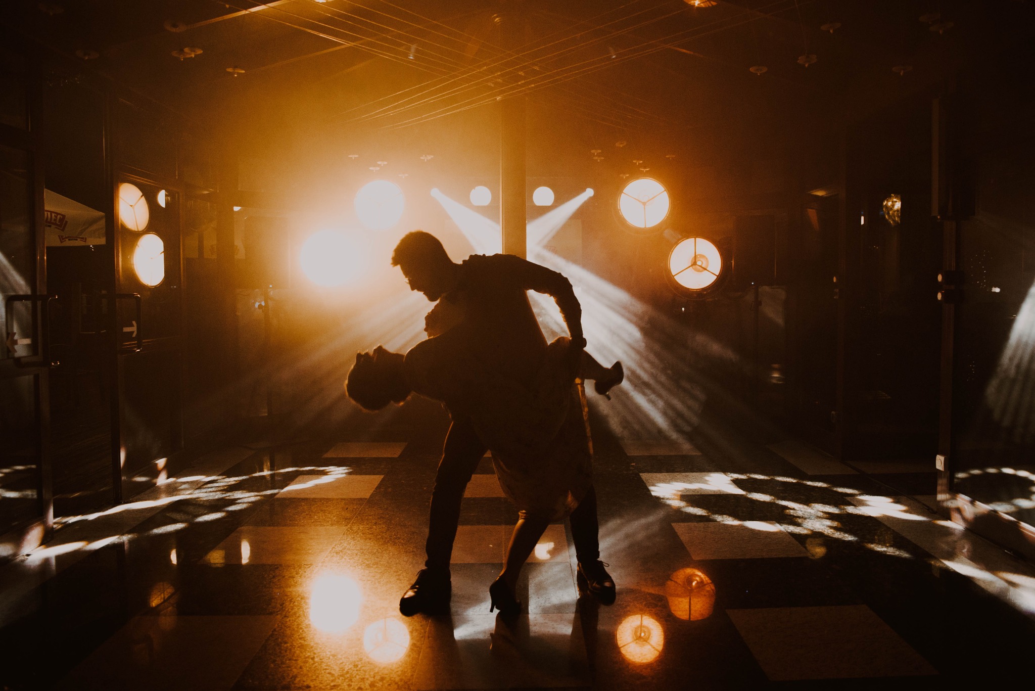 Para taneczna w dynamicznej pozie podczas reportażu ślubnego, silne oświetlenie punktowe tworzy dramatyczne cienie na podłodze.
