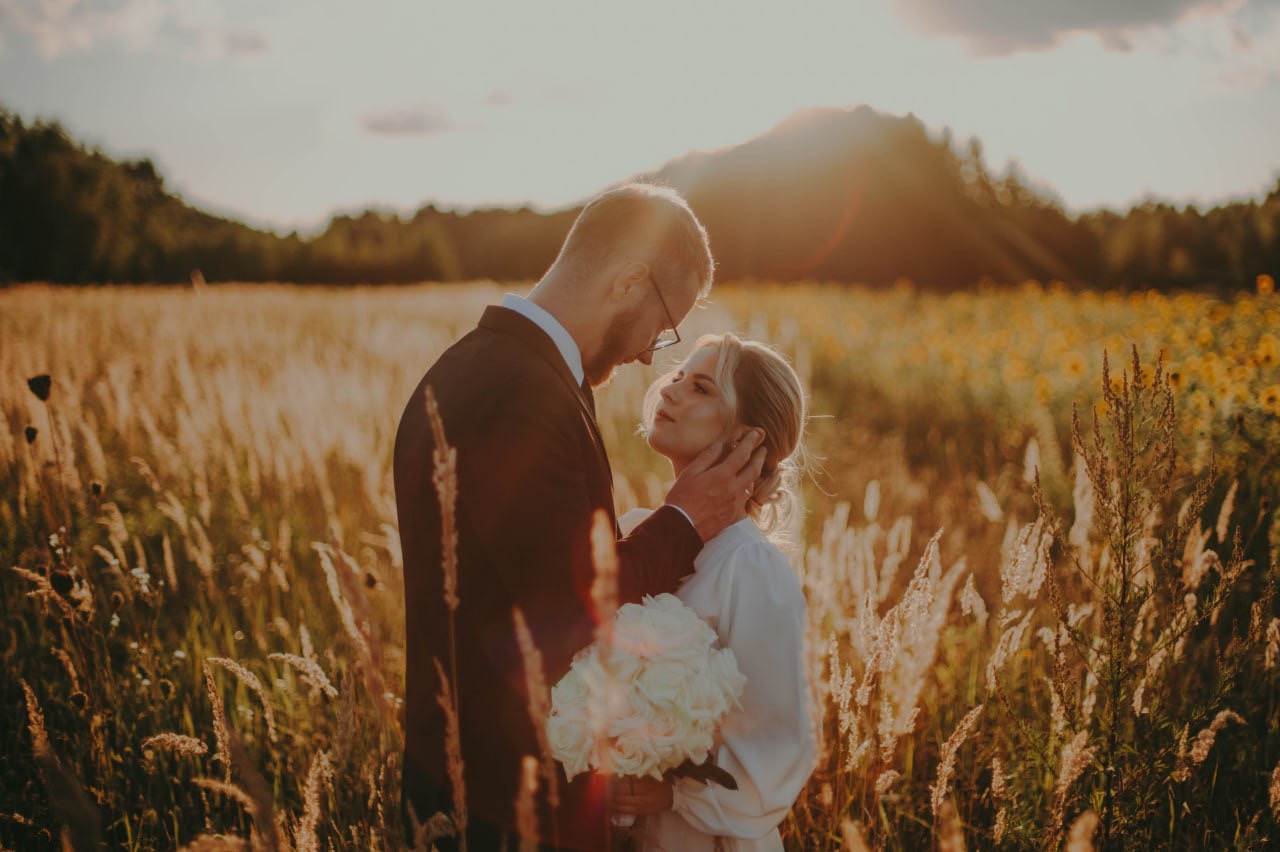 Para młoda w złotym polu trawy podczas zachodu słońca, panna młoda trzyma bukiet białych róż, delikatne dotknięcie dłoni pana młodego na jej policzku.