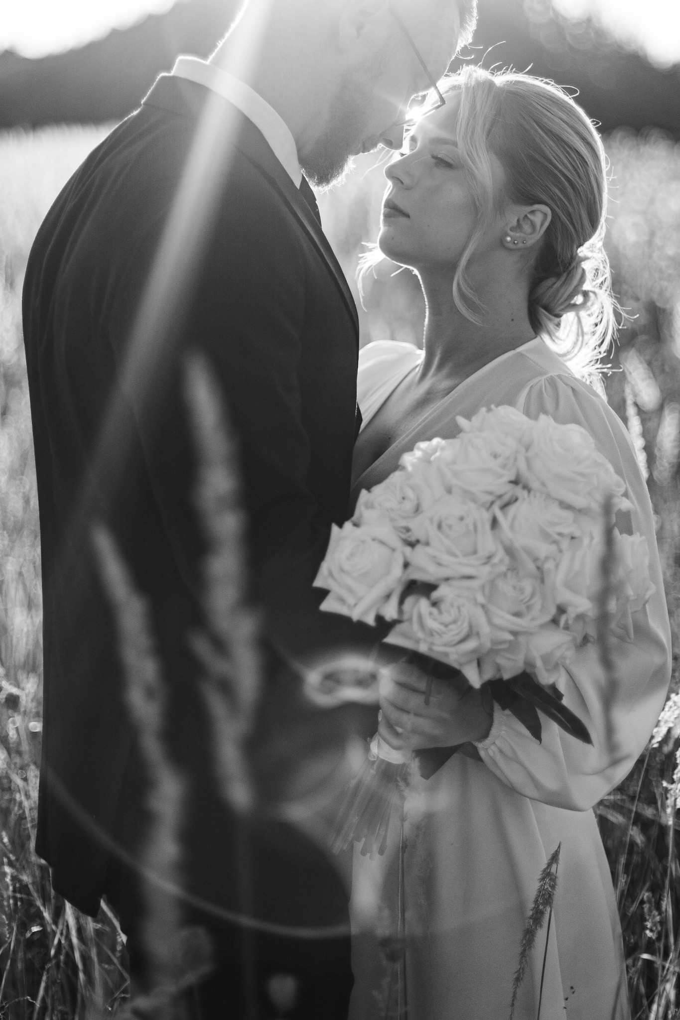 Czarno-białe zdjęcie pary młodej w wysokiej trawie, z promieniami słońca padającymi na twarz kobiety, która trzyma bukiet białych róż.