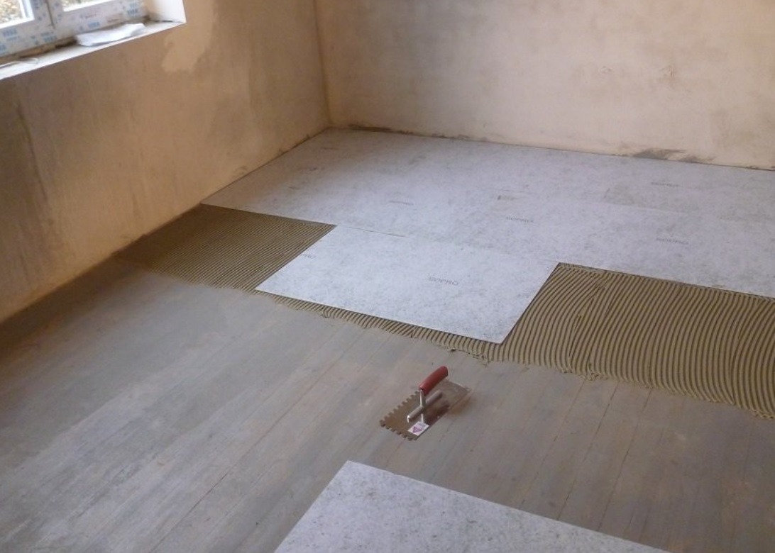 Podłoże pod płytki z naniesionym klejem i częściowo ułożonymi płytami Sopro, widoczna kielnia z czerwoną rączką.
