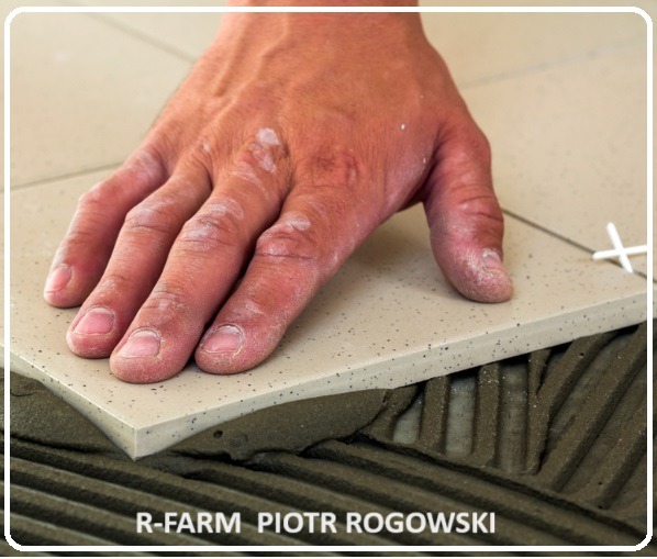 Dłoń dociska beżową płytkę z delikatnym wzorem, osadzoną na warstwie kleju z wyżłobieniami, krzyżyk dystansowy w rogu płytki.