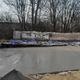 Świeżo wylany, gładki fundament betonowy w wykopie, zabezpieczony niebieską folią, z fragmentami starego muru w tle i nagimi drzewami zimą.