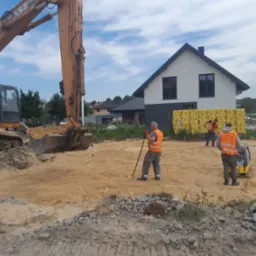 Pomarańczowa koparka CASE na placu budowy, pracownicy w kamizelkach odblaskowych wyrównują teren pod budowę domu jednorodzinnego, w tle widoczne palety z materiałami budowlanymi.