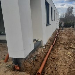 Pietrzak Sp. z o.o. - Brązowe rury ułożone w wykopie wzdłuż ściany budynku, widoczny fragment elewacji z oknami i pracownik w tle, ziemia wokół wykopu nierówna.