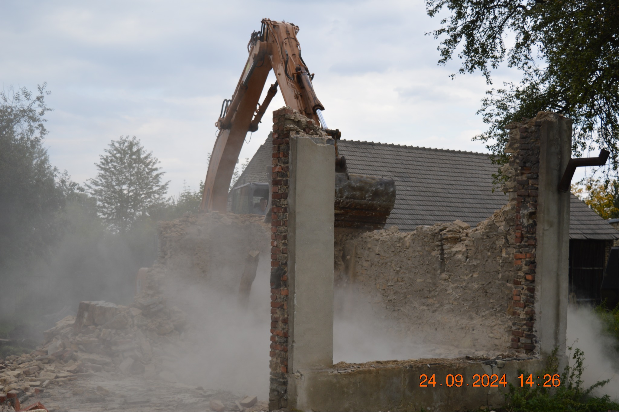Pomarańczowa koparka kruszy pozostałości muru ceglanego podczas rozbiórki budynku, w tle widoczny dach innego budynku i drzewa, unoszący się pył.