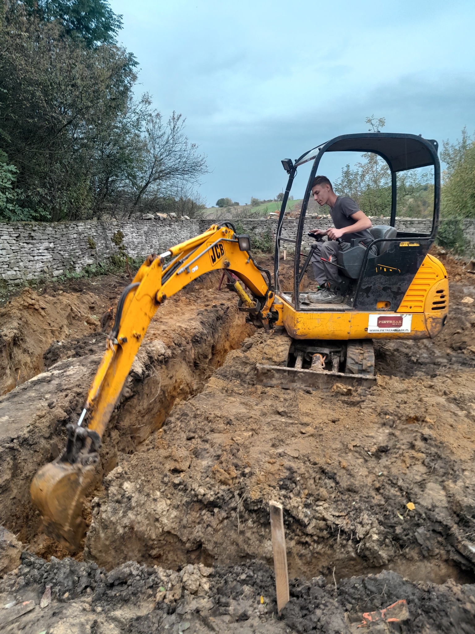 Operator koparki JCB w trakcie wykonywania wąskiego wykopu w ziemi, w tle kamienny murek i drzewa pod pochmurnym niebem.