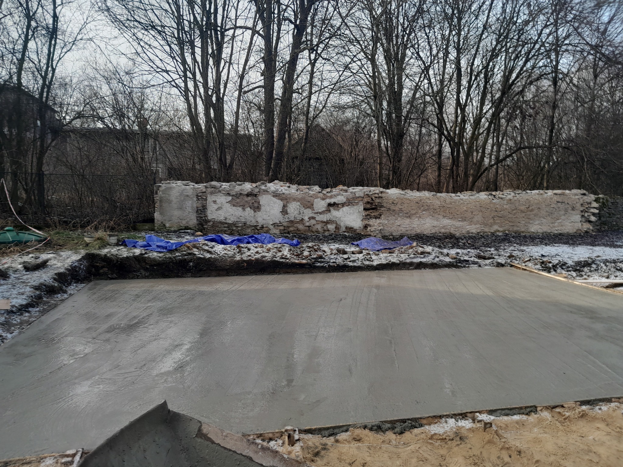 Świeżo wylany, gładki fundament betonowy w wykopie, zabezpieczony niebieską folią, z fragmentami starego muru w tle i nagimi drzewami zimą.
