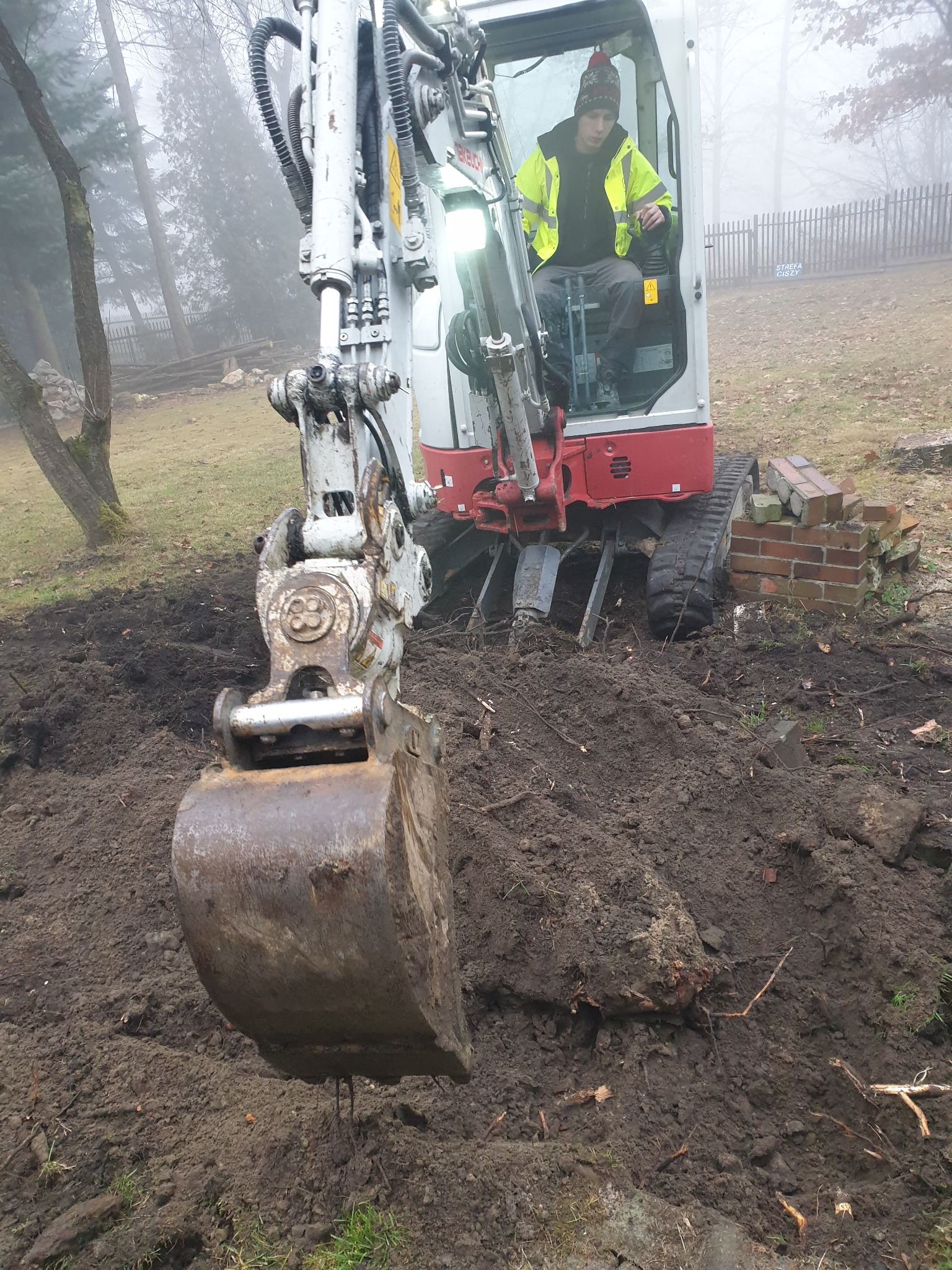 Operator w kabinie małej koparki, łyżka zanurzona w ziemi, obok sterta cegieł, mglisty poranek.