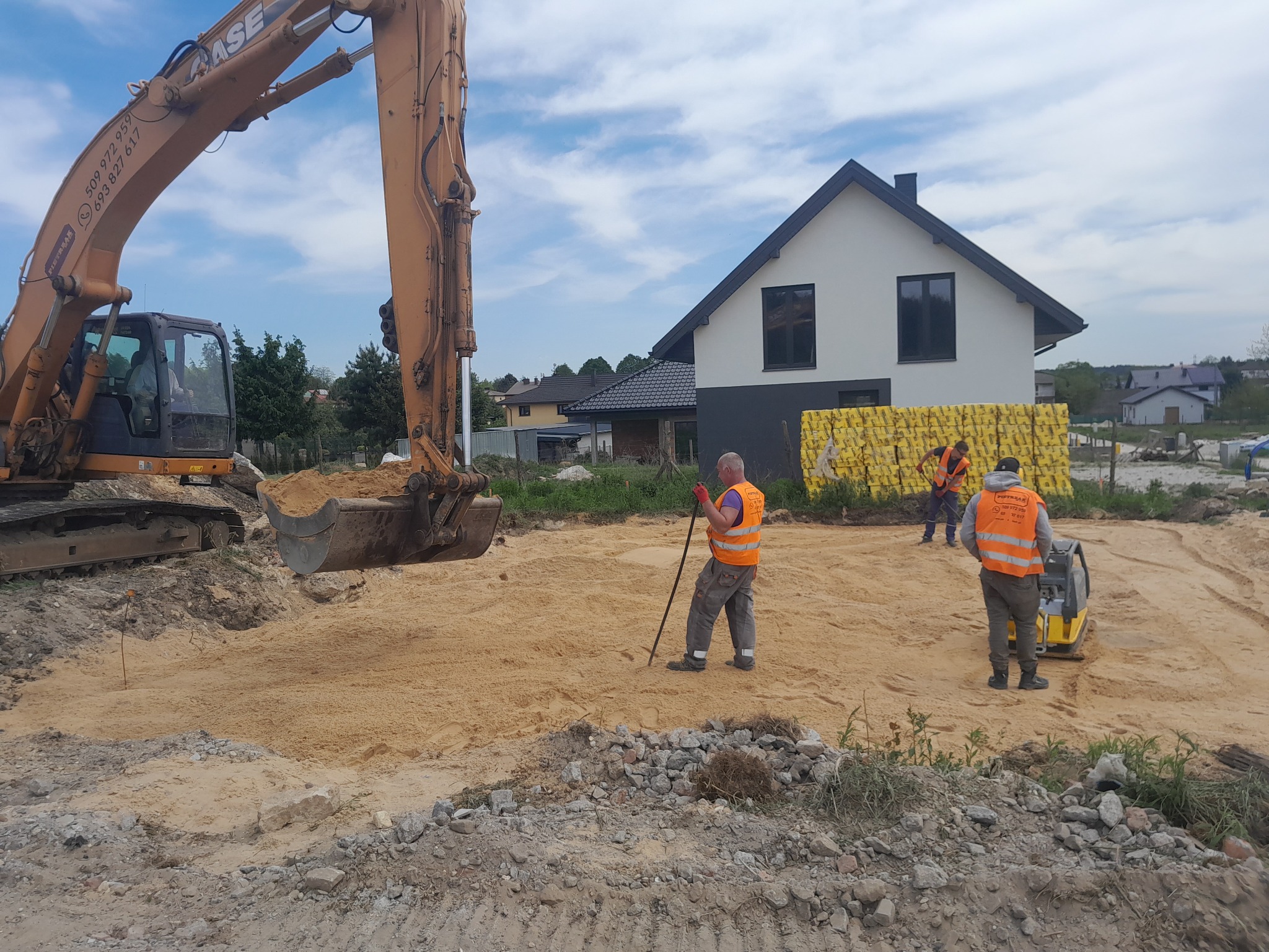 Pomarańczowa koparka CASE na placu budowy, pracownicy w kamizelkach odblaskowych wyrównują teren pod budowę domu jednorodzinnego, w tle widoczne palety z materiałami budowlanymi.