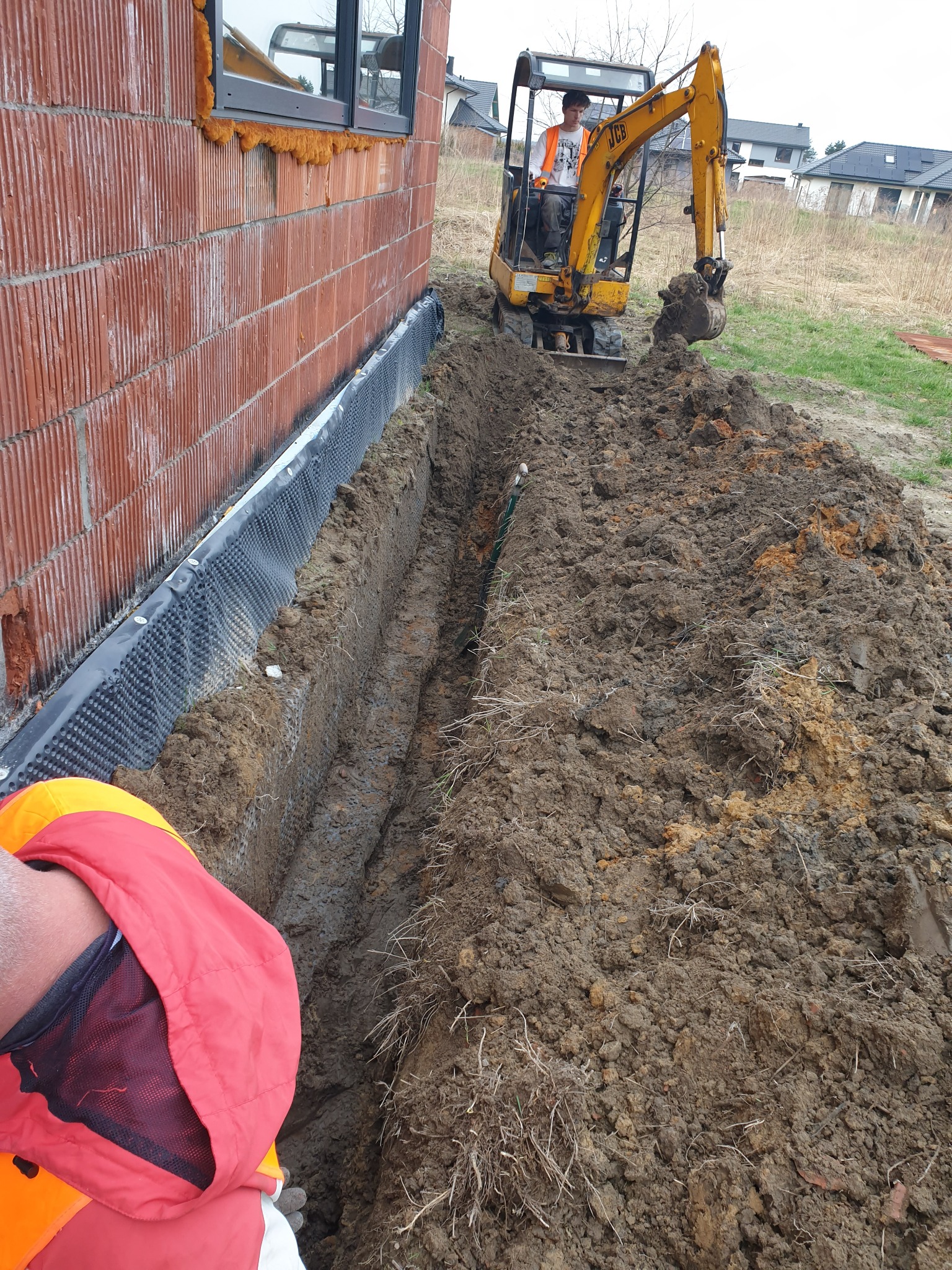 Żółta minikoparka JCB wykonuje wąski wykop wzdłuż ściany budynku z czerwonej cegły z widoczną izolacją przeciwwilgociową, operator w kamizelce odblaskowej.
