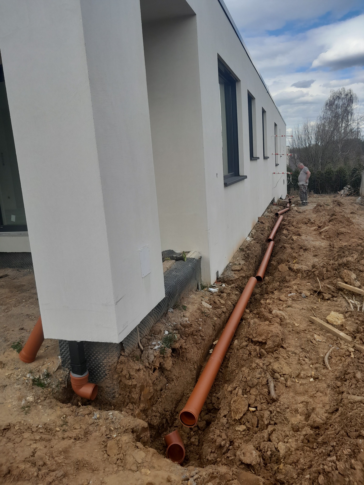 Brązowe rury ułożone w wykopie wzdłuż ściany budynku, widoczny fragment elewacji z oknami i pracownik w tle, ziemia wokół wykopu nierówna.