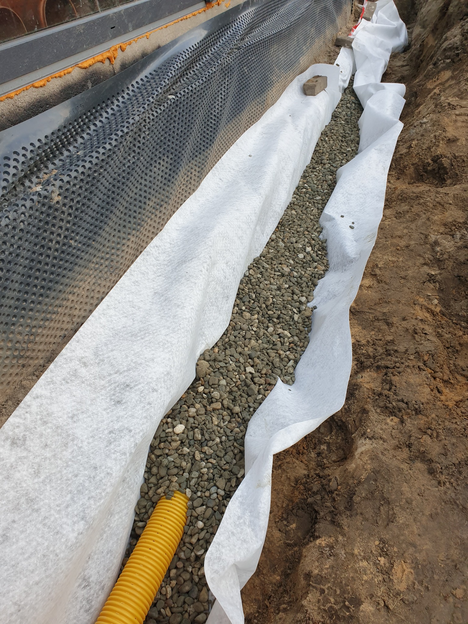 Wykop z żółtą rurą drenażową, obsypany żwirem i wyłożony białą geowłókniną, widoczna czarna folia kubełkowa przy fundamencie.