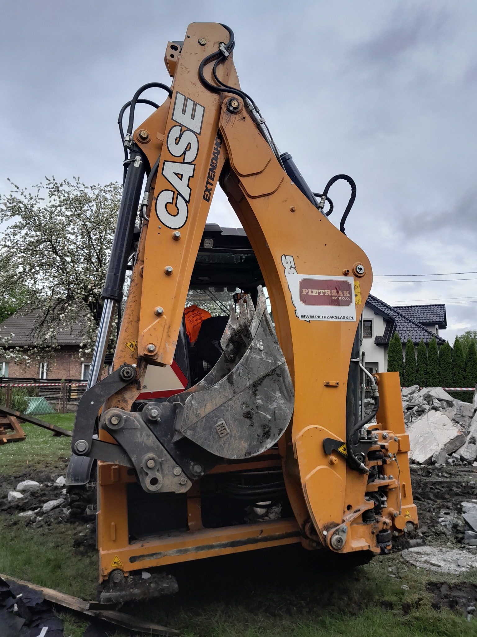 Pomarańczowa koparka Case z łyżką, widoczne gruzy w tle, logo firmy na ramieniu koparki, błotniste podłoże.